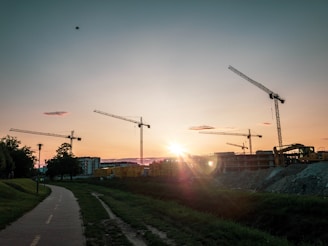 Wide shot of cranes lifting steel beams high above a projeyapı site at sunset.