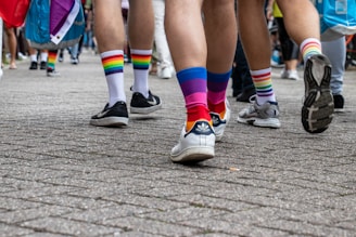 A group of people walking on a cobblestone street, wearing colorful socks and sneakers. The focus is on their legs and feet. Vibrant rainbow patterns on the socks suggest a theme of celebration and unity. The crowd seems to be part of a larger gathering or event.