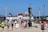 People are walking along a paved path toward a large building by the sea, surrounded by flags from various countries. The sky is clear and blue, suggesting a sunny day. The central focus is a tall clock tower with intricate designs. The area is bustling with tourists dressed in summer attire, indicating a popular seaside destination.