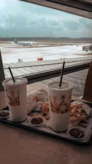 A fast food meal from Burger King is set on a table with a view of an airport runway through a large window. There are two large drink cups with straws, a pile of fries, and packets of ketchup. Outside, an airplane is seen parked on the tarmac under a cloudy sky.