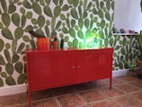 A bright red cabinet stands against a wall covered in a bold green cactus pattern. On top of the cabinet, there are several potted cacti of varying shapes and sizes. To the right, a shelf displays colorful ornamental skulls. The room has a lively and eclectic decor with terra-cotta tiled flooring.