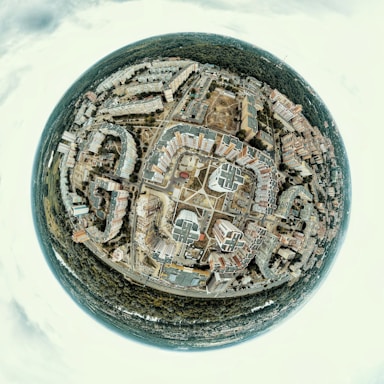 A spherical panorama capturing a cityscape with numerous buildings arranged in a circular pattern around a central square. The image includes residential and commercial buildings, roads intersecting at various points, and a dense forest surrounding the urban area.