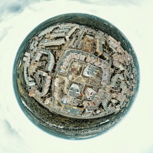A spherical panorama capturing a cityscape with numerous buildings arranged in a circular pattern around a central square. The image includes residential and commercial buildings, roads intersecting at various points, and a dense forest surrounding the urban area.