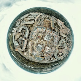 A spherical panorama capturing a cityscape with numerous buildings arranged in a circular pattern around a central square. The image includes residential and commercial buildings, roads intersecting at various points, and a dense forest surrounding the urban area.