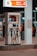 A fuel pump station at night illuminated by artificial lights. The pump displays options for diesel and premium gas, with hoses neatly hung. Signs around the pump provide safety instructions and branding. The background shows a few people and additional pump stations.