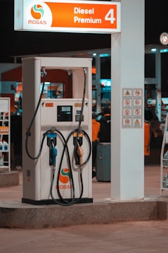 A fuel pump station at night illuminated by artificial lights. The pump displays options for diesel and premium gas, with hoses neatly hung. Signs around the pump provide safety instructions and branding. The background shows a few people and additional pump stations.