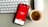 A smartphone displaying a red screen with the text indicating a subscriber count of 164,913 is placed on a gray surface. Nearby, a white keyboard and a white mouse can be seen along with a cup of coffee with frothy cream.