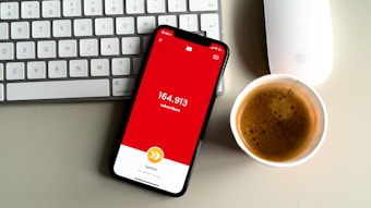 A smartphone displaying a red screen with the text indicating a subscriber count of 164,913 is placed on a gray surface. Nearby, a white keyboard and a white mouse can be seen along with a cup of coffee with frothy cream.
