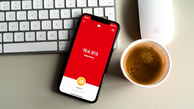 A smartphone displaying a red screen with the text indicating a subscriber count of 164,913 is placed on a gray surface. Nearby, a white keyboard and a white mouse can be seen along with a cup of coffee with frothy cream.