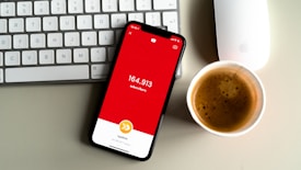 A smartphone displaying a red screen with the text indicating a subscriber count of 164,913 is placed on a gray surface. Nearby, a white keyboard and a white mouse can be seen along with a cup of coffee with frothy cream.