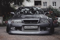 A sleek, modified sports car with a lowered stance is parked on a textured pavement. The car features a customized front bumper and pronounced headlights, giving it a distinctive, aggressive look. The license plate indicates a Russian origin. Surrounding the car are hints of greenery, and a building facade is visible in the background.