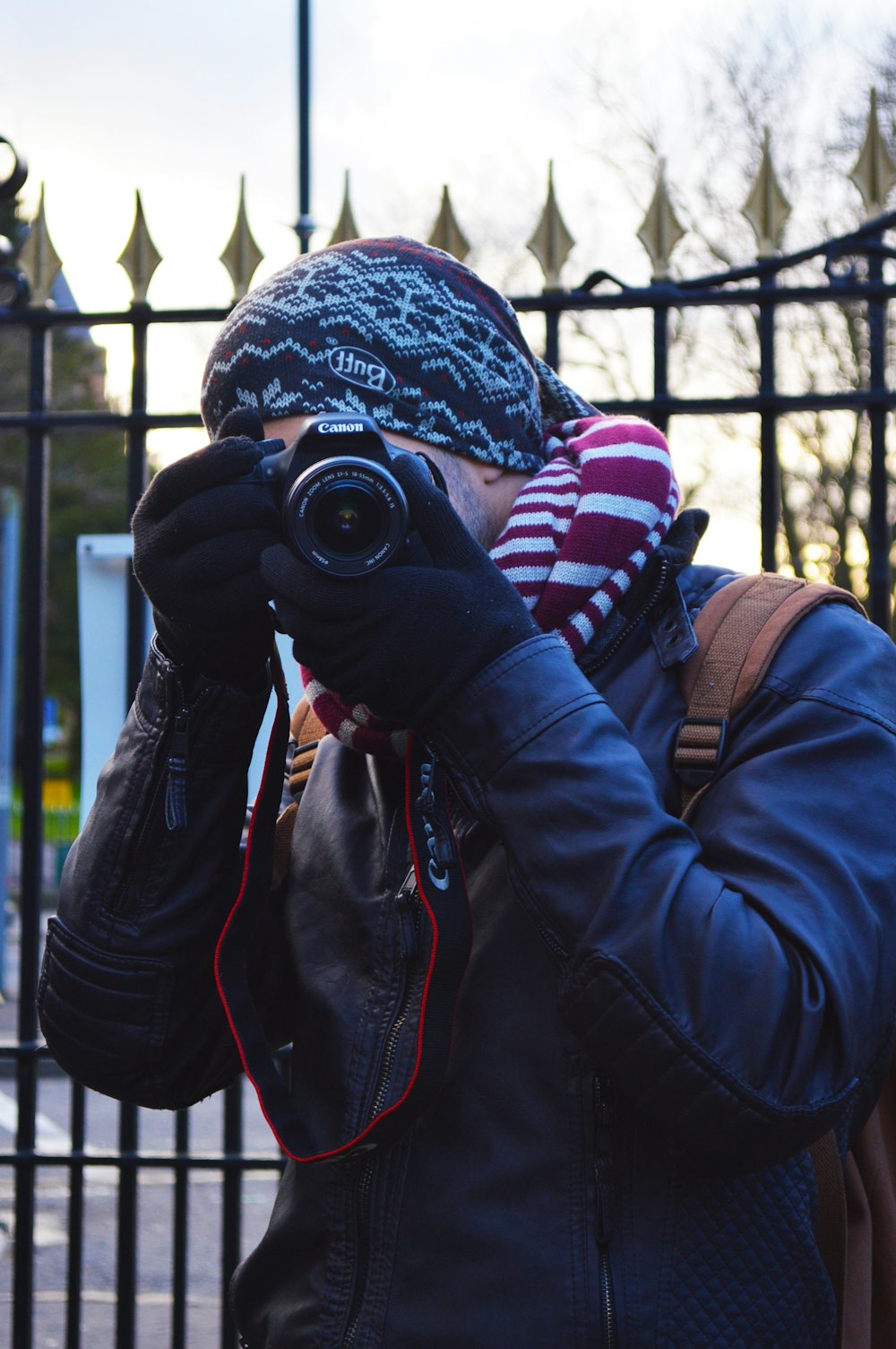 man wearing black jacket using black Canon DSLR camera near black metal gate