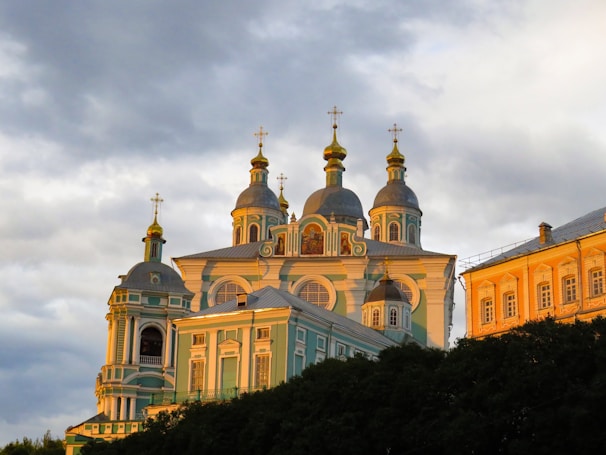 A beautifully designed Orthodox church with multiple domes topped with golden crosses, bathed in the warm light of the setting sun. The building features intricate architectural details and a light blue facade with white accents. Adjacent to the church is another ornate building with a golden-yellow exterior.