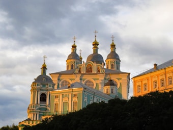 A beautifully designed Orthodox church with multiple domes topped with golden crosses, bathed in the warm light of the setting sun. The building features intricate architectural details and a light blue facade with white accents. Adjacent to the church is another ornate building with a golden-yellow exterior.