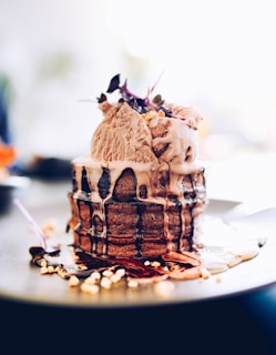 A stack of chocolate pancakes topped with scoops of ice cream and garnished with small edible flowers sits on a plate. Chocolate syrup is drizzled over the pancakes and ice cream, pooling around the base along with sprinkled nuts.