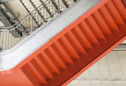 A steel staircase in an industrial setting, featuring bright orange steps and a light gray railing with visible bolts and beams. Overhead metal structures and a concrete ceiling complement the industrial aesthetic.