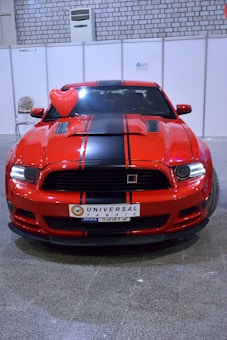 A shiny red sports car with black racing stripes is parked indoors. The license plate indicates a location, and there is a red heart-shaped balloon attached to the driver's side mirror. The background consists of a tiled wall and a white exhibition panel.