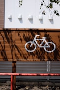 A brown wooden fence displays a painted white bicycle symbol, indicating a parking or designated cycling area. The background consists of a white wall with four small white lights evenly spaced. Shadows of leaves from a nearby tree cast intricate patterns across the fence and the wall, adding a natural touch. A red metal barrier runs horizontally along the bottom of the image, hinting at a boundary or protective feature.