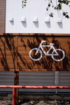 A brown wooden fence displays a painted white bicycle symbol, indicating a parking or designated cycling area. The background consists of a white wall with four small white lights evenly spaced. Shadows of leaves from a nearby tree cast intricate patterns across the fence and the wall, adding a natural touch. A red metal barrier runs horizontally along the bottom of the image, hinting at a boundary or protective feature.