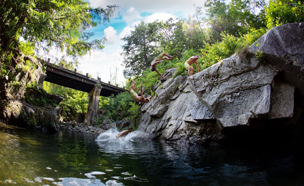 four men about to jump on lake surrounded with tall and green trees