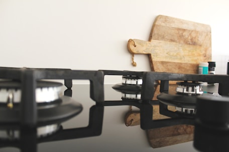 A modern kitchen stove top with three gas burners is visible, featuring black metal supports and reflective glass surface. In the background, there is a wooden cutting board with a handle, leaning against a light-colored wall. On the right side, several spice containers add a touch of color.