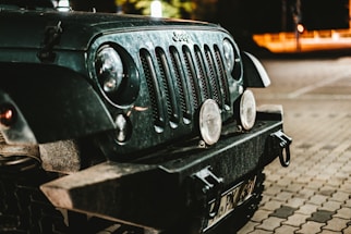 The front end of a rugged off-road vehicle is captured in a dimly lit environment. The vehicle features round headlights, a distinctive grille, and appears to be dusty, suggesting recent use in dusty or off-road conditions. The background shows part of a parking area, subtly illuminated by streetlights, providing a nighttime atmosphere.
