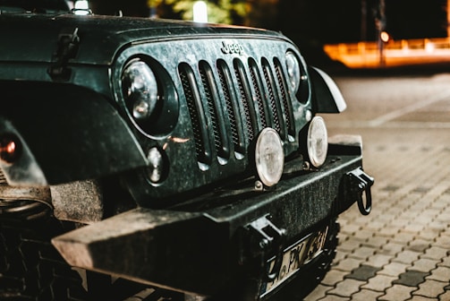 The front end of a rugged off-road vehicle is captured in a dimly lit environment. The vehicle features round headlights, a distinctive grille, and appears to be dusty, suggesting recent use in dusty or off-road conditions. The background shows part of a parking area, subtly illuminated by streetlights, providing a nighttime atmosphere.