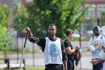 A group of young men wearing sports gear are preparing for a lacrosse game outdoors. One person in the foreground holds a lacrosse stick and wears a protective glove and a sleeveless jersey with a logo. In the background, others are seen holding lacrosse sticks, one of whom is wearing a helmet.