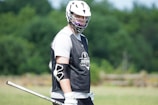 A lacrosse player wearing a black mesh vest with the logo 'Jaguars Lacrosse' is standing on a grassy field. He is wearing a white helmet and a black arm guard, holding a lacrosse stick. The background features green trees blurred out of focus.