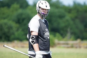 A lacrosse player wearing a black mesh vest with the logo 'Jaguars Lacrosse' is standing on a grassy field. He is wearing a white helmet and a black arm guard, holding a lacrosse stick. The background features green trees blurred out of focus.