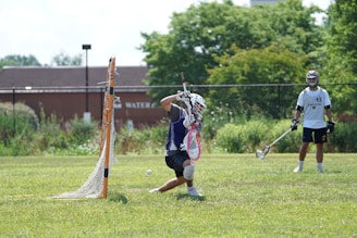 A lacrosse player in protective gear is actively defending the goal with a stick, while another player stands nearby holding their own lacrosse stick. The setting is a grassy field surrounded by lush greenery, and a building and fence are visible in the background.