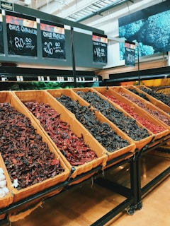 Various types of dried chilies are arranged in large, rectangular bins within a grocery store. The chilies vary in color, including dark red and black, and are accompanied by signs displaying their respective prices. The overhead lighting gives the area a warm hue.