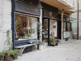 A quaint flower shop with a rustic exterior features a variety of green plants in both pots and hanging baskets. The shop front is adorned with large windows displaying an assortment of floral arrangements and a wooden bench with more plant pots. The signage above the window displays a minimalistic design, and ivy climbs along the wall, adding to the cozy, inviting atmosphere of the street corner setting.