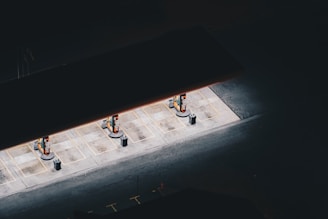 Aerial view of a dimly-lit gas station with three fuel pumps, isolated in darkness. The concrete ground is highlighted by overhead lights, creating a stark contrast with the surrounding shadows.