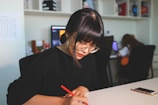 A person with glasses is seated at a table, focused on writing with a red pencil. In the background, a computer screen and another individual are visible. The setting appears to be an office or study space with shelves containing various objects.