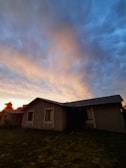 A single-story house with a dark exterior is silhouetted against a vibrant sunset sky filled with dramatic clouds. The sky transitions from deep blue to soft oranges and pinks, casting a warm glow over the scene.