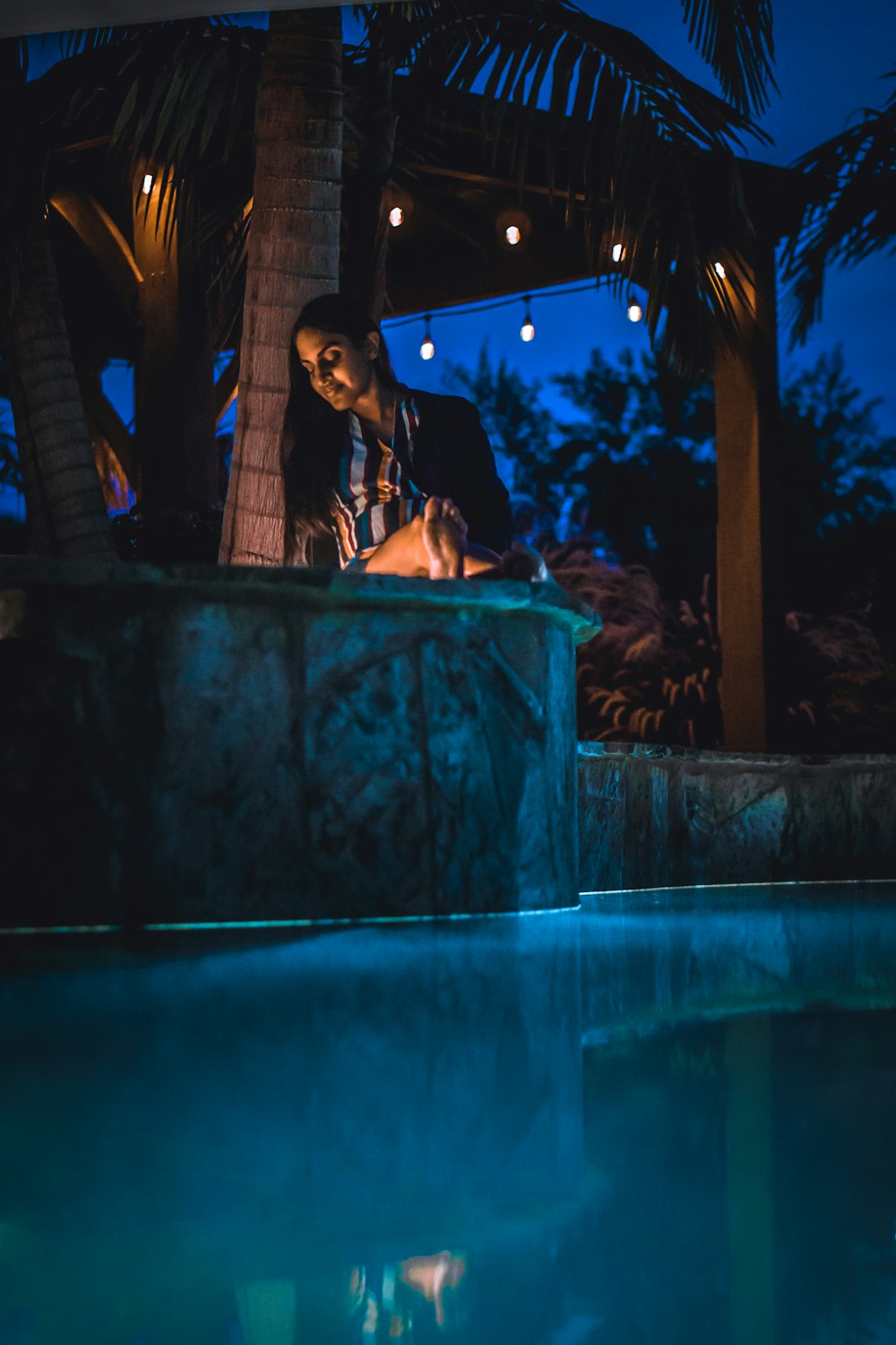 夜間、照明のついたペンダント電球のそばのプールに座っている女性