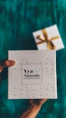 A decorative white box with small star patterns is being held by a pair of hands. The box is labeled 'Vya Naturals' with a note indicating it is free from parabens and sulphates. In the background, there is another similarly styled box with a golden ribbon, slightly out of focus. The surface beneath is a rich teal color.