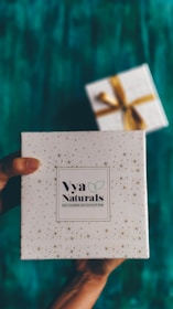 A decorative white box with small star patterns is being held by a pair of hands. The box is labeled 'Vya Naturals' with a note indicating it is free from parabens and sulphates. In the background, there is another similarly styled box with a golden ribbon, slightly out of focus. The surface beneath is a rich teal color.