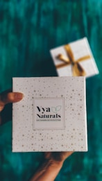 A decorative white box with small star patterns is being held by a pair of hands. The box is labeled 'Vya Naturals' with a note indicating it is free from parabens and sulphates. In the background, there is another similarly styled box with a golden ribbon, slightly out of focus. The surface beneath is a rich teal color.