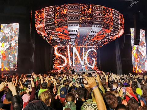 A large crowd of people at a concert, with a stage featuring a massive, illuminated sign that reads 'SING'. The stage setup includes vibrant lights and screens displaying colorful, abstract visuals. Many attendees are holding up smartphones to capture the event.