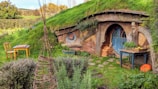 A charming, rustic house built into a grassy hillside features a round blue door with circular windows. The roof and walls are covered with green grass, blending the structure into the natural landscape. A small wooden table with a chair stands nearby, and a table with fruits and plants rests to the side. There is a woven wooden structure in the foreground, and an orange pumpkin on the ground adds a pop of color.