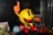 A colorful figurine of Pac-Man is displayed, characterized by a bright yellow body, red boots, and orange gloves. The character is depicted pointing upward with one hand while standing on a surface resembling a maze. Surrounding it are various themed memorabilia, including game elements like cherries and ghost figures in blue and red. Comic books and other related decorations are also visible.