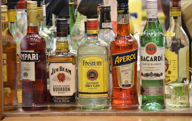 A collection of various liquor bottles is displayed on a shelf. The bottles include brands like Campari, Jim Beam, Finsbury, Aperol, and Bacardi. Each bottle is distinct in shape and color, with labels showcasing their logos and brand names.