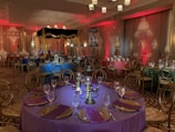 An elegantly decorated banquet hall with round tables draped in rich-colored tablecloths, including purple, blue, and green. Each table is set with glasses, plates, and napkins, featuring ornate centerpieces. The room is dimly lit with ambient lighting in red, contributing to an intimate atmosphere. Chandeliers hang from the ceiling, and decorative lanterns and drapes enhance the setting.