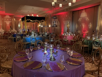 An elegantly decorated banquet hall with round tables draped in rich-colored tablecloths, including purple, blue, and green. Each table is set with glasses, plates, and napkins, featuring ornate centerpieces. The room is dimly lit with ambient lighting in red, contributing to an intimate atmosphere. Chandeliers hang from the ceiling, and decorative lanterns and drapes enhance the setting.