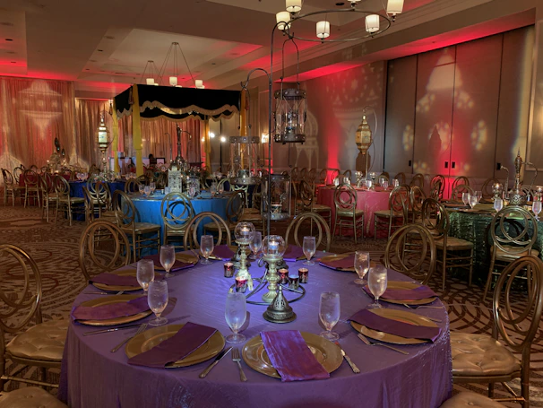 Elegant banquet tables dressed in white linens with purple napkins and soft pink lighting for an evening event.