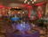 An elegantly decorated banquet hall with round tables draped in rich-colored tablecloths, including purple, blue, and green. Each table is set with glasses, plates, and napkins, featuring ornate centerpieces. The room is dimly lit with ambient lighting in red, contributing to an intimate atmosphere. Chandeliers hang from the ceiling, and decorative lanterns and drapes enhance the setting.