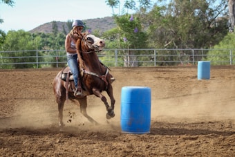 A person wearing a helmet and casual clothing is riding a brown horse. The horse is in motion, appearing to make a quick turn around a blue barrel on a dirt surface. There is a fenced area in the background, surrounded by trees and greenery with hills visible in the distance.