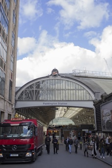 An entrance to Paddington Station is visible, featuring a large arched glass roof with intricate design. A red delivery truck is parked at the forefront, and a group of people, including commuters and pedestrians, are walking in and out of the station. The weather is clear with blue skies and scattered clouds. Tall buildings are seen on the left side.